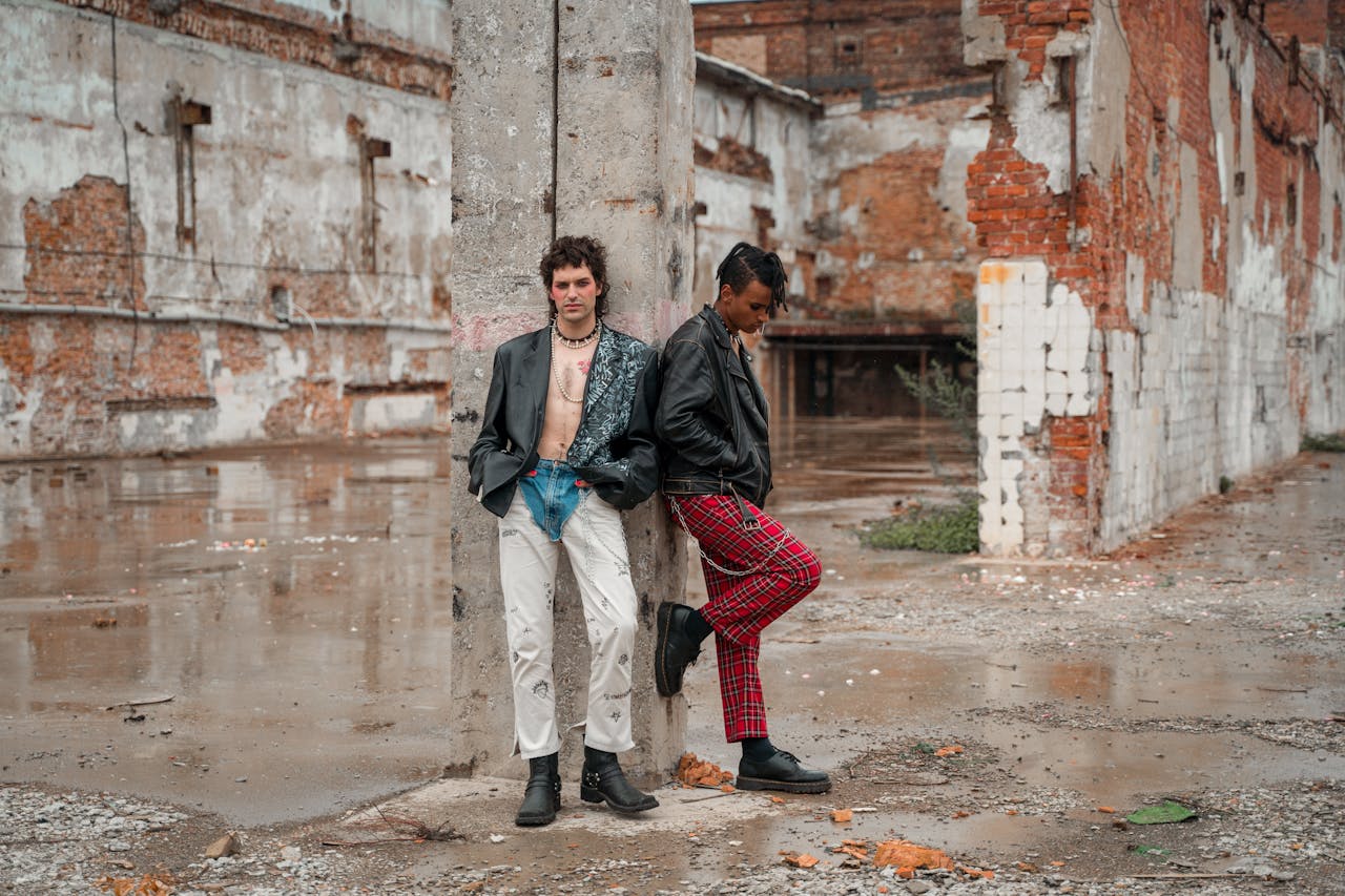 Two male models in leather jackets pose stylishly at an abandoned industrial site, showcasing bold streetwear fashion.