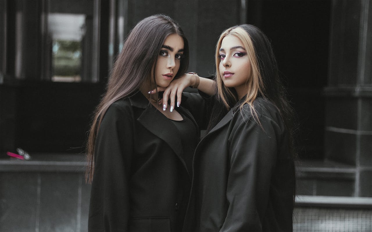 Two fashionable women in black coats pose with confidence against a dark, urban backdrop. Modern and chic look.